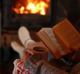 Woman resting with cup of hot drink and book near fireplace